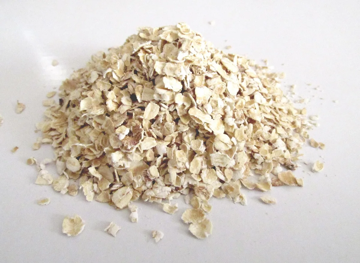 Hands pouring rolled oats into a measuring cup on a bright kitchen counter with a jar of nuts and a sliced apple nearby