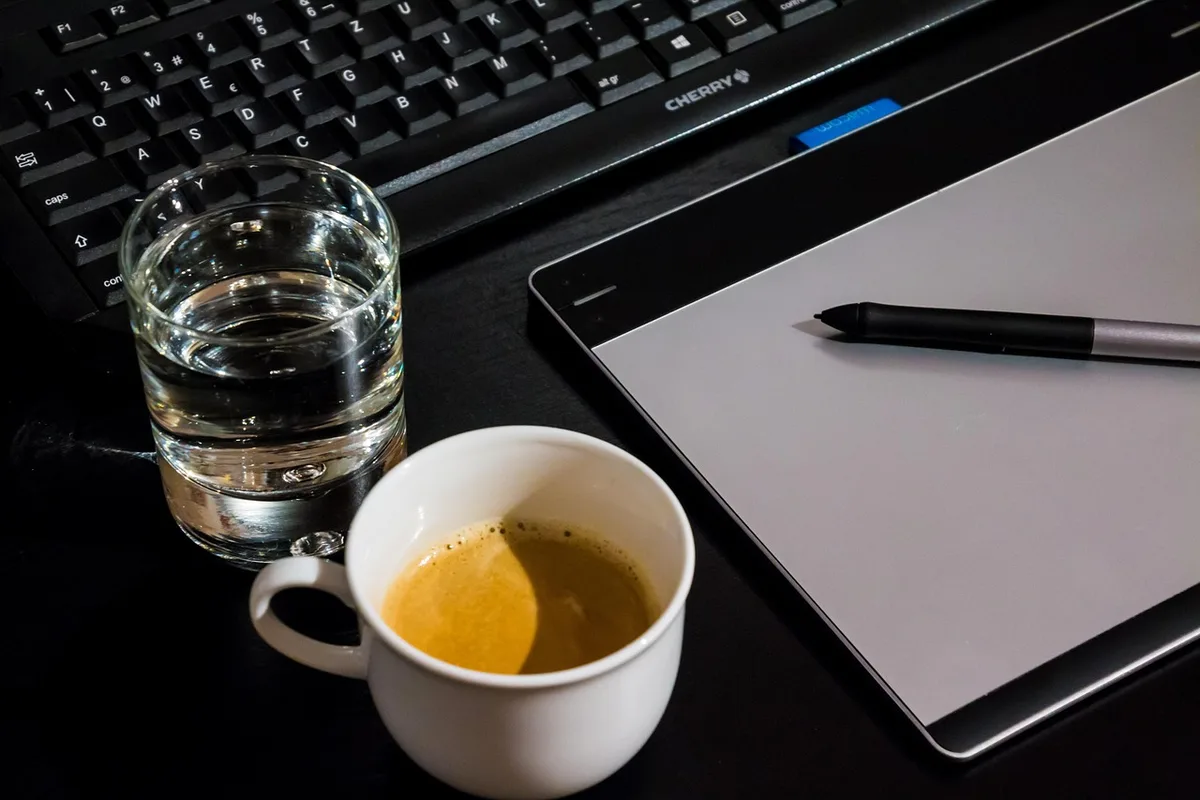A neatly arranged desk with hands holding a tablet displaying a magazine table of contents, a notepad and pen beside it, a mug and reading glasses nearby